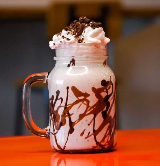 Milkshake in a mason jar with whipped cream and chocolate drizzle on an orange table.